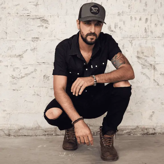 Bold black man with a beard crouching against a white brick wall, casual style, promoting hope and faith.