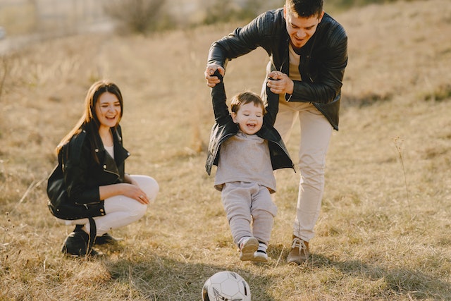 A mom and dad helping their son kick a soccerball on the field.