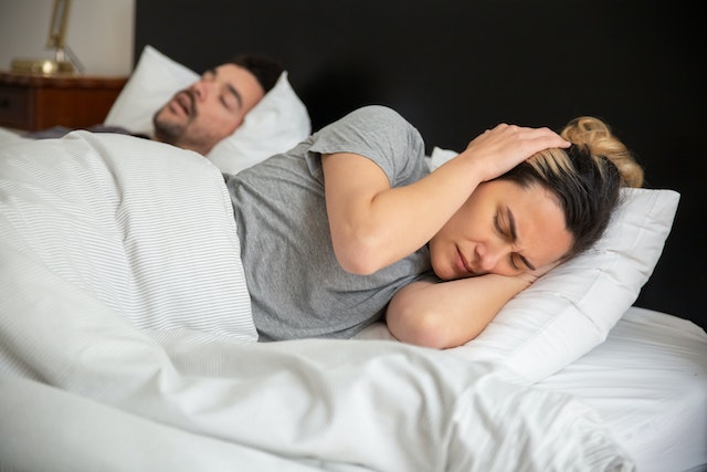 Two people laying in bed. The man is snoring and the lady is covering her ears.